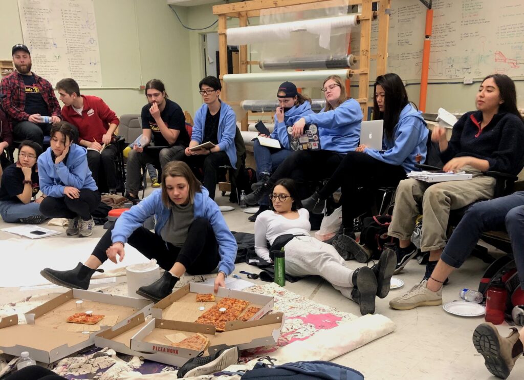 People sitting on the floor and chairs of a room, looking at notes and eating pizza.