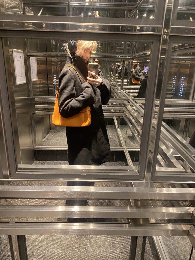 Woman takes mirror selfie in elevator while holding orange bag.