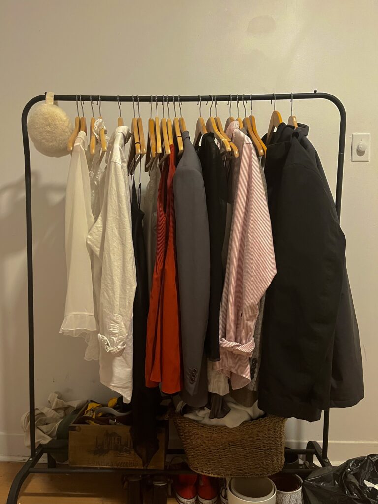 Items of clothing hang on wooden hangers on a black clothing rack against a white wall.