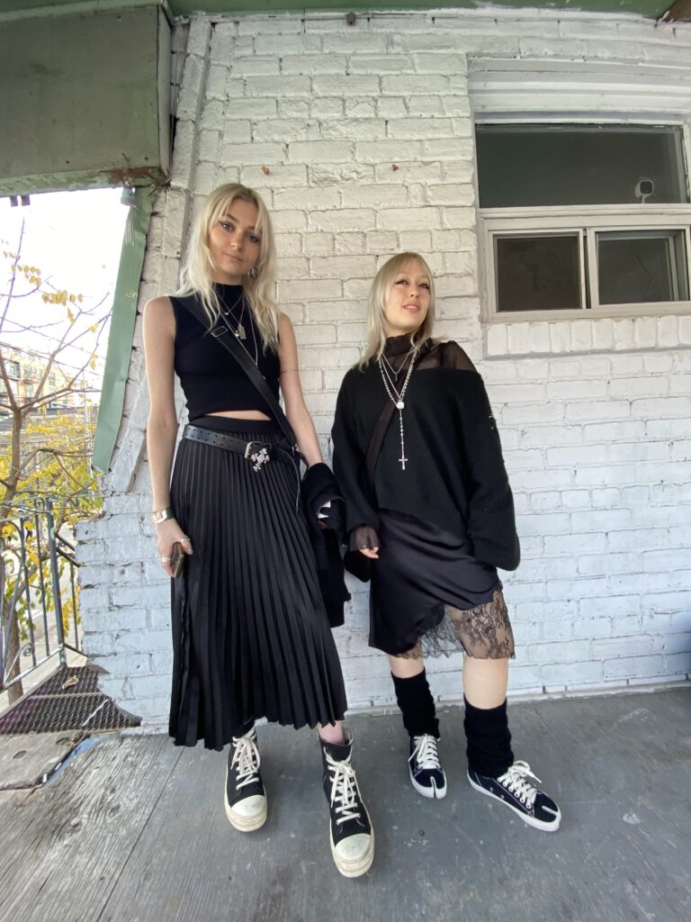 Two young women in all black outfits and silver jewelry pose for a photo against a white brick wall.